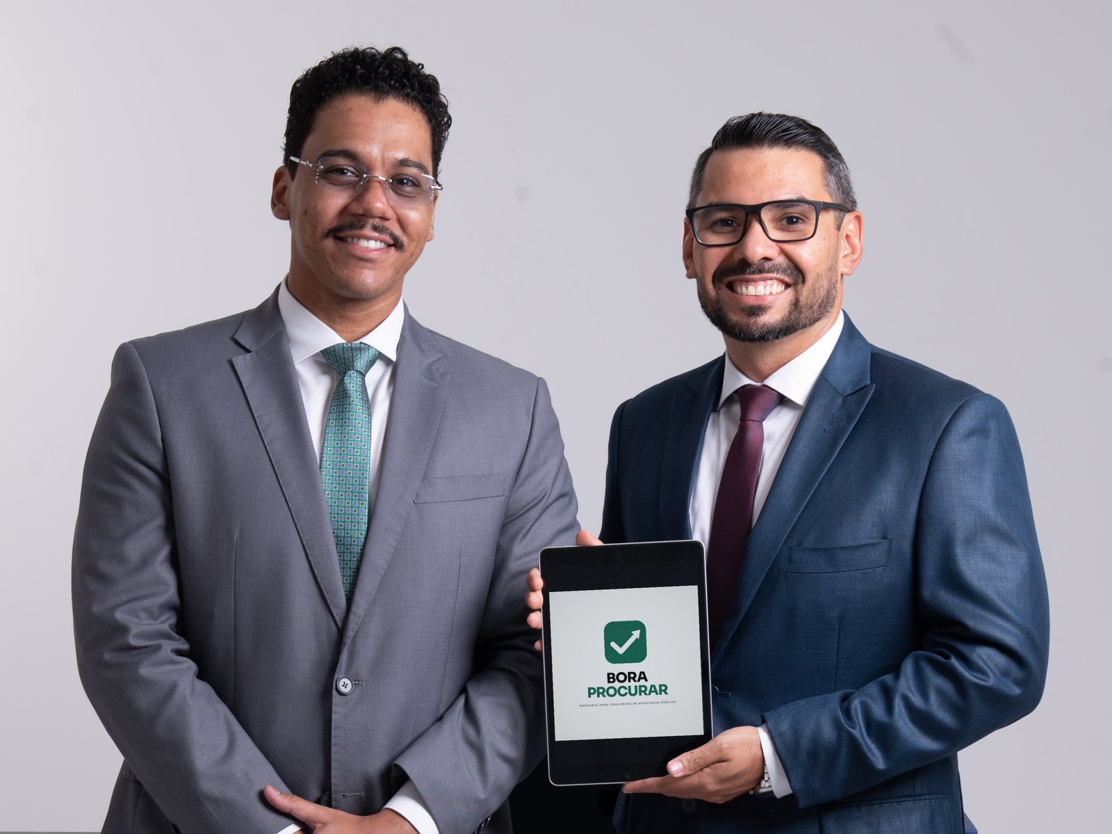 Os mentores André Casas e Juan Desidério, de terno, com um tablet que mostra a marca Bora Procurar